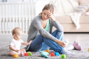 A mother who looks sad is sitting with her baby on the floor with toys all around.