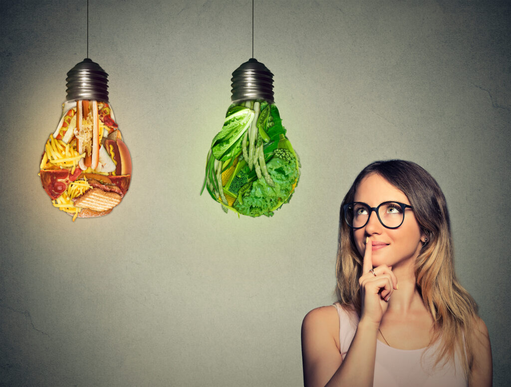 Portrait beautiful woman in glasses thinking looking up at junk food and green vegetables shaped as light bulb isolated on gray background. Diet choice right nutrition healthy lifestyle concept